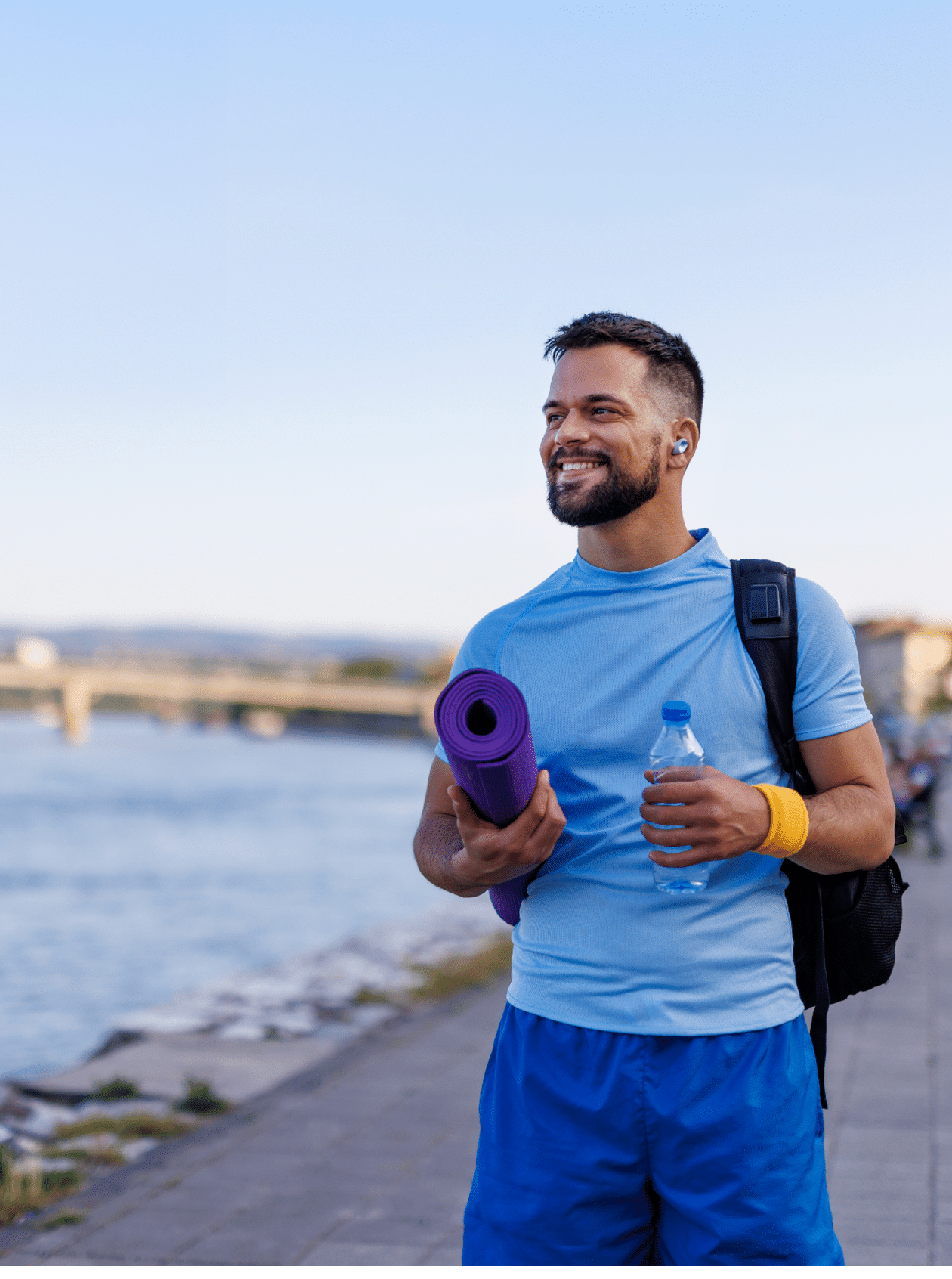 Man stands by the riverside, ready for a workout with a rolled-up yoga mat, water bottle, and a backpack. Dressed in a blue shirt and yellow wristband, he looks invigorated.